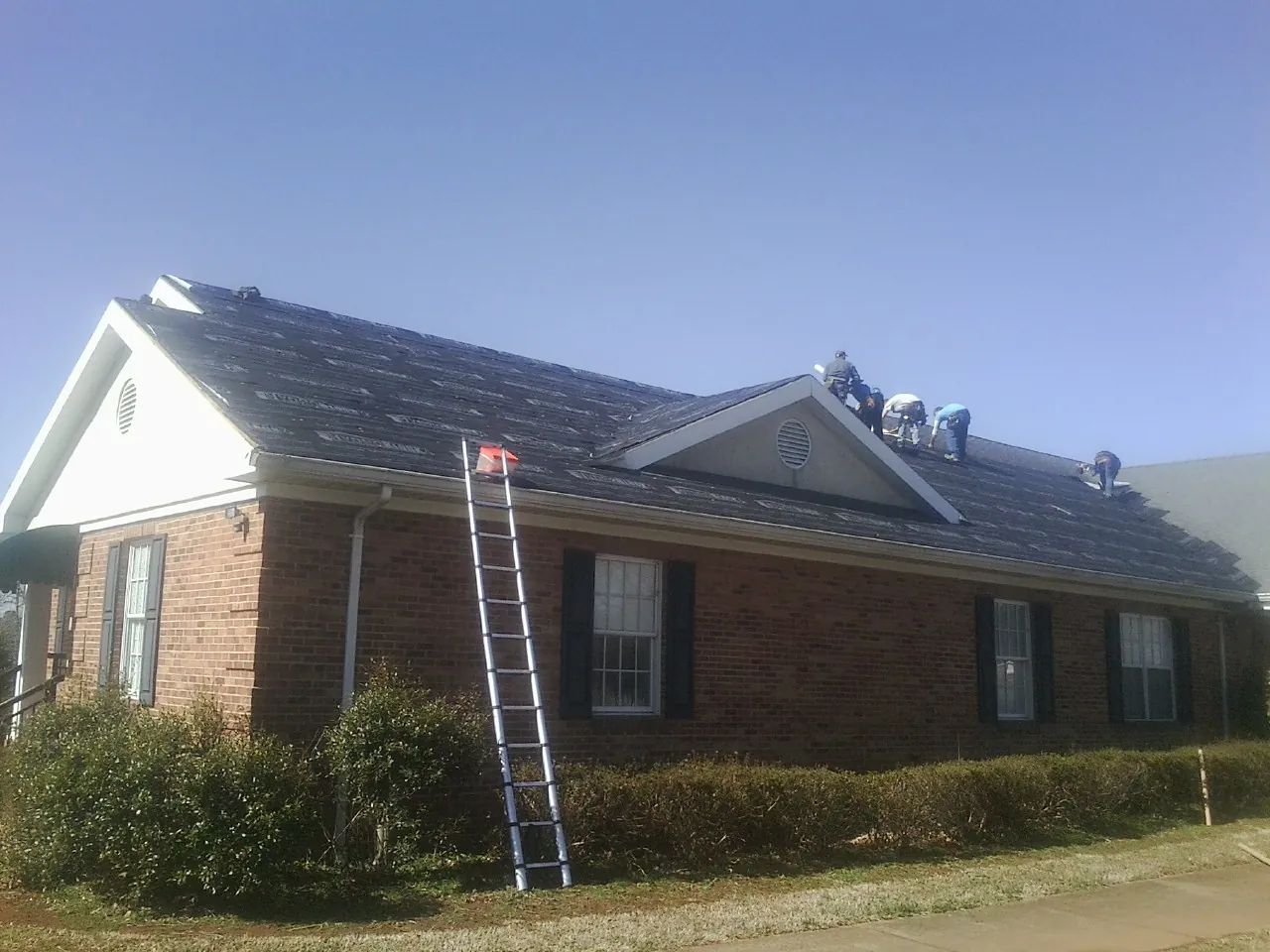 Crew tearing shingles off a brick ranch roof with a ladder set — Metal Roof Repair work by our crew in Morrisonville