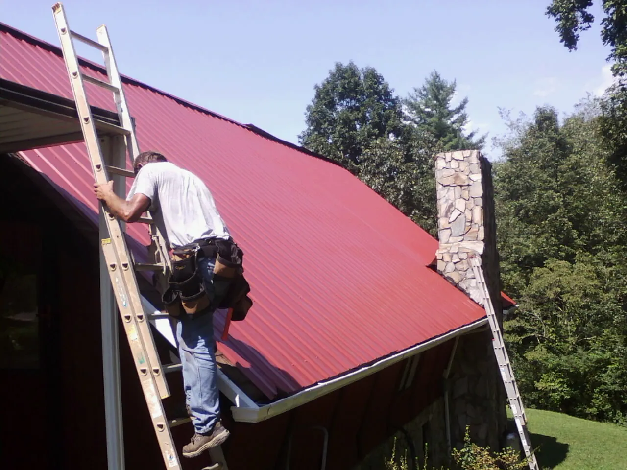Roofer on a ladder beside a red metal roof and stone chimney — Soffit & Fascia Repair by Roofing Brothers in Morrisonville
